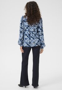 Blå mönstrad blus med långa ärmar, som har en geometrisk design och gula accenter, ihop med mörk denim byxor med utsvängda ben.