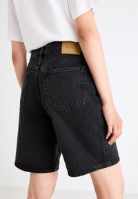 Black denim shorts with a straight cut, featuring two back pockets and a tan leather patch on the waistband. Smooth texture, casual design.