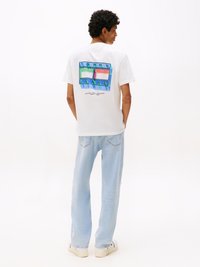Valkoinen puuvillateeppari, jossa on värikäs graafinen printti selässä ja tekstillä "TOMMY JEANS", yhdistettynä vaaleansinisiin farkkuihin.