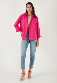 Fuchsia oversized button-up shirt with rolled sleeves, paired with light blue cropped jeans and multicolored strappy heels.