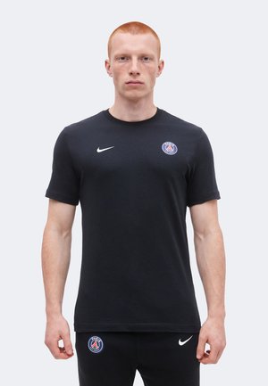 Jeune homme aux cheveux roux courts portant un t-shirt de sport noir Nike Paris Saint-Germain et un pantalon noir assorti, faisant face.