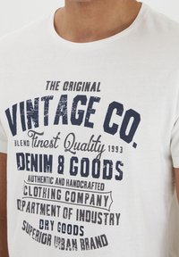 Bijela pamučna majica s mornarskom grafikom, uključujući "VINTAGE CO." i razne detalje brenda, otisnuta u različitim stilovima fonta.