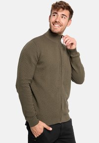 Olivengrøn zip-up sweater med et struktureret chevronmønster, ribbet krave og manchetter samt en tætsiddende pasform. Synlig lynlås og håndplacering på lommen.