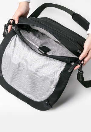 Schwarze Umhängetasche aus glattem Leder, mit einem Reißverschlussverschluss und einem grau gemusterten Innenfutter, das über ein kleines Fach und einen Stoffgurt verfügt.