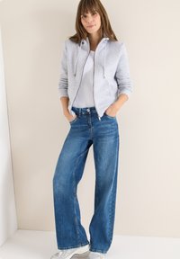 Šedá mikina na zip s tkaničkami, nošená přes bílou košili, spojená s širokými modrými džíny a stříbrnými teniskami. Neutrální pozadí.