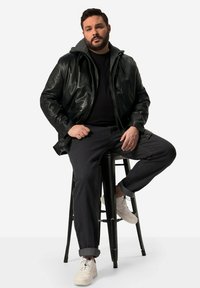 Giacca di pelle nera con cappuccio grigio, pantaloni scuri e sneakers bianche. La figura è seduta su uno sgabello nero con uno sfondo neutro.