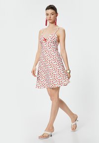 Witte zomerjurk met rode aardbeiprint, fijne verstelbare schouderbandjes en een aansluitend lijfje. Uitlopende rok en een klein rood strikdetail aan de voorkant.