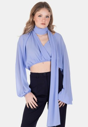 Giovane donna con capelli mossi sciolti che indossa un top corto azzurro maniche lunghe con sciarpa attaccata e pantaloni neri a vita alta.