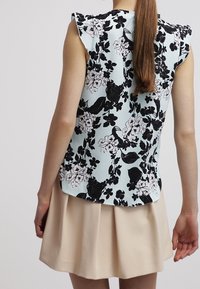 Donna con capelli lunghi che indossa un top senza maniche di colore blu con una stampa floreale in bianco e nero e una gonna plissettata beige, vista da dietro.