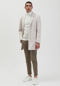 Licht beige overjas met een notch-kraag, gedragen over een witte coltrui, gecombineerd met een bruine geruite broek en witte sneakers.