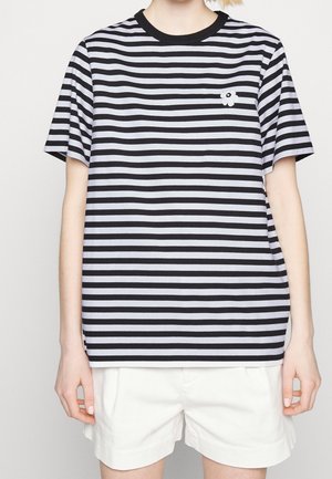 Person trägt ein schwarz-weiß horizontal gestreiftes T-Shirt mit einem kleinen Blumendetail, kombiniert mit weißen Shorts.