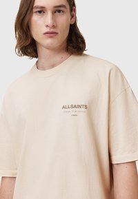 Béžové oversized bavlněné tričko s kulatým výstřihem a krátkými rukávy. Na přední straně, nad lemem, je potisk s logem v tmavě hnědé barvě.