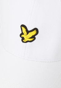 Casquette blanche en tissu texturé avec un emblème d'aigle noir et jaune sur le côté. Le design est simple et sportif.