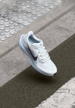 Weißer Nike-Laufschuh mit schwarzem Swoosh, der über Asphalt nahe einem Betonbordstein und tastbarem Pflaster auf dem Gehweg schwebt.