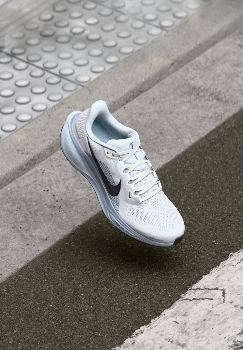 Chaussure de running Nike blanche avec un swoosh noir flottant au-dessus de l'asphalte près du bord en béton et du pavage tactile sur le trottoir.