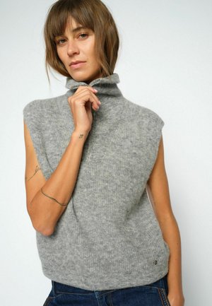 Kvinde iført ærmeløs grå strikket rullekravesweater, som trækker kraven op, med tatoveringer på armene, mod en ensfarvet lys baggrund.