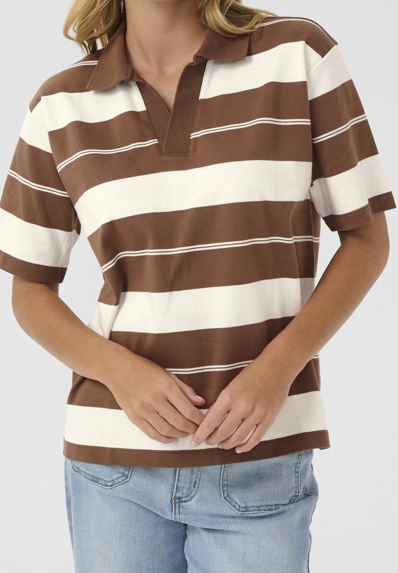 Femme portant un polo à manches courtes larges rayures marron et blanc, et un jean bleu clair, les mains doucement jointes devant.