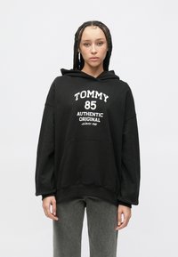 Čierna mikina s kapucňou vyrobená z jemnej látky, s bielym nápisom "TOMMY 85 AUTHENTIC ORIGINAL" a predným vreckom. Voľný strih s rebrovanými manžetami.