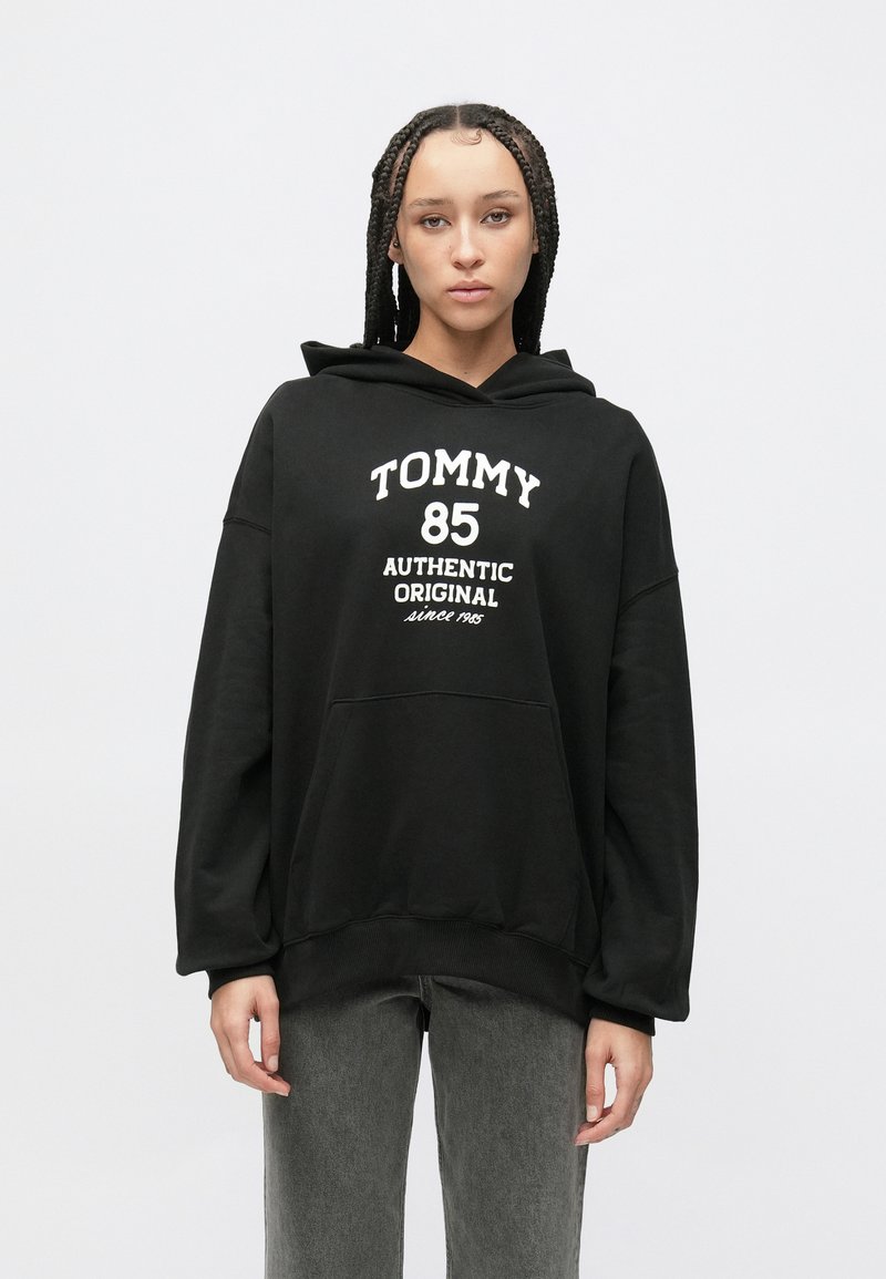 Čierna mikina s kapucňou vyrobená z jemnej látky, s bielym nápisom "TOMMY 85 AUTHENTIC ORIGINAL" a predným vreckom. Voľný strih s rebrovanými manžetami.