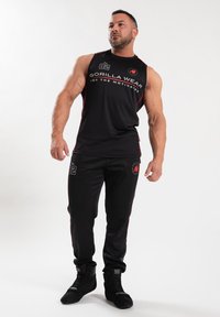 Zwarte sportieve tanktop met rode accenten en logo, gecombineerd met zwarte joggers met rode zijstripes en logo. Geweven stof en aangesloten design.