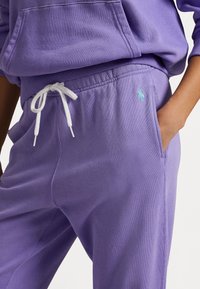 Lila mjukisbyxor i mjukt bomull, med dragsko i midjan med vita snören och en liten blå logga i vänstra fickområdet.