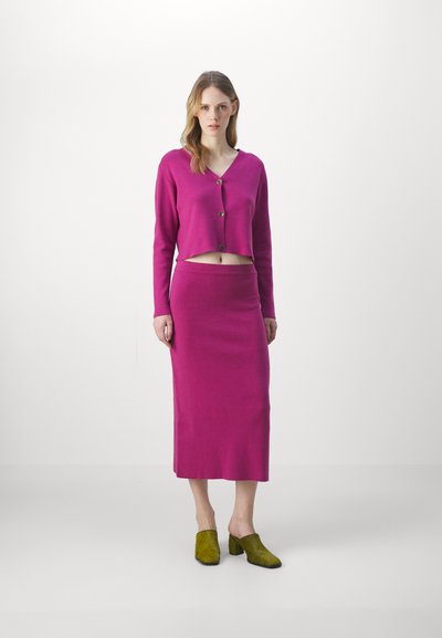 Fuchsia verkürzter Cardigan mit Knöpfen, kombiniert mit einem fuchsia-farbenen Midirock. Das Modell trägt grüne, strukturierte Mules. Weicher Stoff, figurbetonter Schnitt.