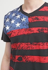 Svart T-shirt med en slitna amerikansk flagga-design i blått, rött och vitt, V-ringning och korta ärmar.
