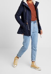 Mörkblå parka med en mjuk crèmefärgad foder, med en justerbar huva, knapprar och kombinerad med ljusblå jeans och beige höga sneakers.