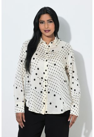 Femme aux longs cheveux noirs portant un chemisier blanc à pois noirs et un pantalon noir, debout devant un fond blanc uni.