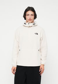 Giovane che indossa un pullover in pile color crema con tasche frontali e cappuccio, abbinato a pantaloni neri, in piedi contro uno sfondo bianco.