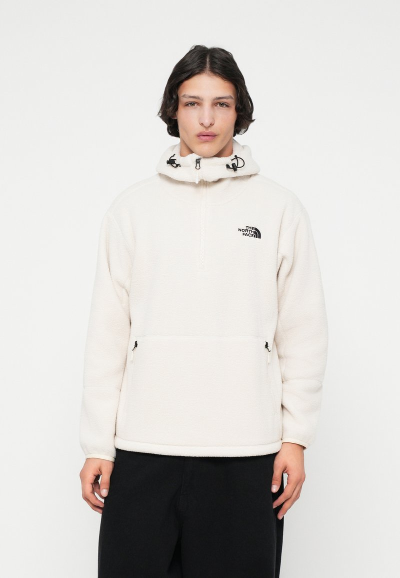 Giovane che indossa un pullover in pile color crema con tasche frontali e cappuccio, abbinato a pantaloni neri, in piedi contro uno sfondo bianco.