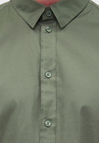 Olivově zelená košile na knoflíky s klasickým límečkem a šesti knoflíky. Hladká textura s jemným šitím a čistým, přiléhavým střihem.