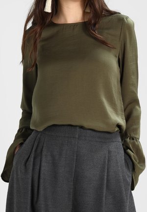 Femme portant un chemisier vert olive à manches longues avec des poignets évasés et un pantalon plissé gris taille haute, les mains dans les poches.