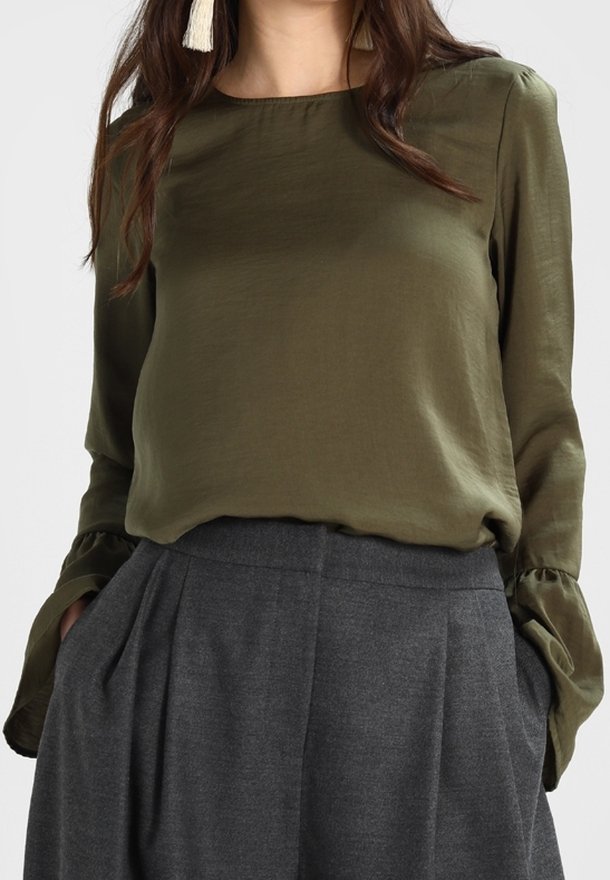 Femme portant un chemisier vert olive à manches longues avec des poignets évasés et un pantalon plissé gris taille haute, les mains dans les poches.