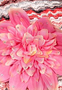 Roze stofbloem met gelaagde bloemblaadjes, versierd met rhinestones, op een gestructureerde, multikleurige achtergrond met zigzagpatronen.