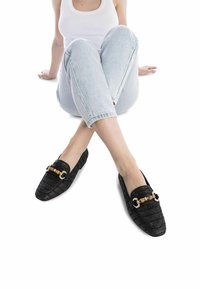 Mocasines negros trenzados con una tela texturizada, punta cuadrada, acento de cadena en dorado y negro, combinados con jeans claros de corte enrollado.
