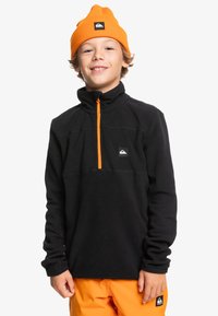 Schwarzer Fleece-Pullover mit hohem Kragen, orangefarbenem Reißverschluss und Logo-Patch. Getragen mit einer orangefarbenen Mütze und passenden Hosen. Weicher Stoff.