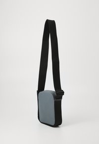 Šedá látková ramenní taška čtvercového tvaru, s černým popruhem a minimalistickým designem. Disponuje zapínáním na zip a polstrovanou texturou.