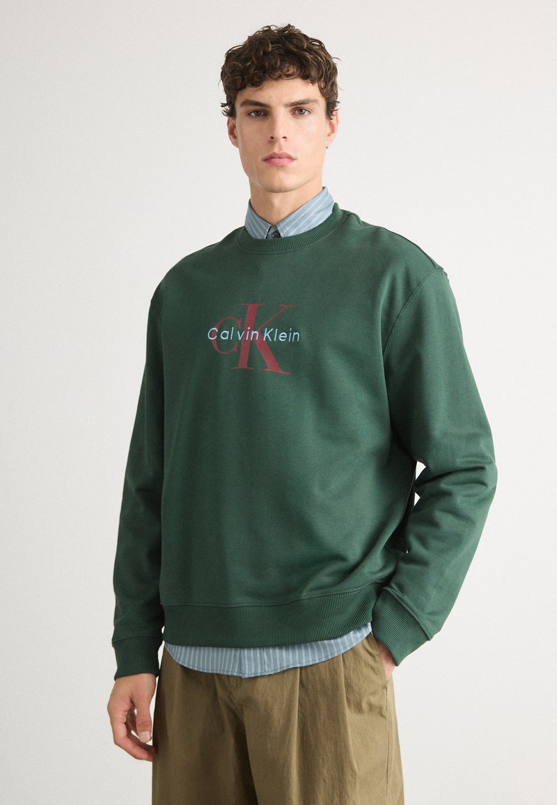 Sweatshirt verde com decote redondo, destacando o texto "Calvin Klein" em vermelho e azul. Usado sobre uma camisa listrada azul clara, combinado com calças cáqui.