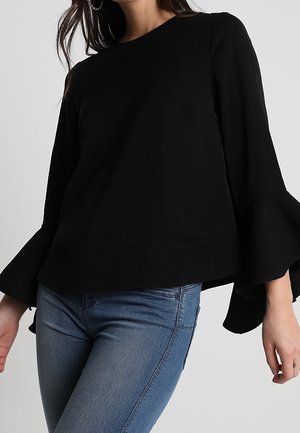 Vrouw draagt een zwarte longsleeve met gerimpelde trompetmouwen en blauwe jeans, staand tegen een effen grijze achtergrond.