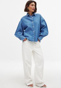 Denim button-up shirt in light blue with oversized design, featuring a collar and large buttons, paired with high-waisted white wide-leg pants.