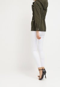 Oliwkowa kurtka z kapturem ze ściągaczem w talii, dwoma bocznymi kieszeniami i srebrnymi suwakami. Połączona z białymi obcisłymi spodniami i czarnymi szpilkami.