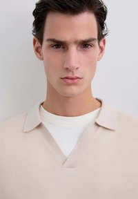 Pull en V beige avec une texture douce et une chemise intérieure blanche contrastante, dotée d'un col rabattu et d'un design épuré.