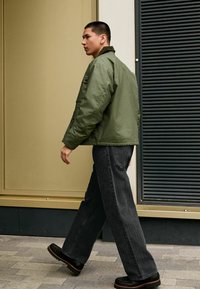 Grüne Bomberjacke mit cropped Design, großen Taschen, kombiniert mit lockeren schwarzen Jeans und schwarzen Schuhen. Steht auf einem Steinpflaster.