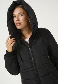 Veste rembourrée noire avec capuche, fermeture éclair et poignets élastiques. Elle présente un design matelassé et des cordons de réglage sur la capuche.