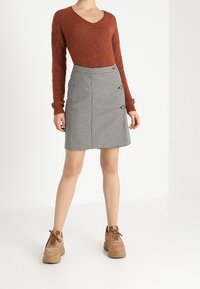 Un pull tricoté de couleur rouille avec un col en V, associé à une jupe en pied-de-poule grise et noire comportant trois boutons décoratifs. Des baskets marron.