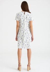 Frau steht und trägt ein knielanges weißes Kleid mit blauem Blumenmuster und kurzen Ärmeln, kombiniert mit beigen High-Heels.
