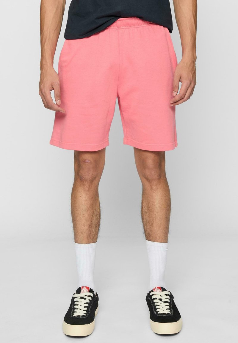 Herre rosa bomuldsshorts med elastisk talje, længde over knæet, og sidelommer, parret med sorte sneakers og hvide sokker.