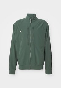 Mørkegrøn zip-up jakke med ståkrave, to lodrette brystlynlåse, sidelommer, sorte striber på ærmerne og lille hvid Nike-logo på fronten.