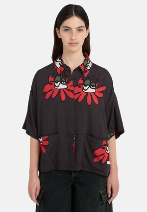 Nő laza fekete gombos ingben, amelyen piros és zöld rajzfilmszerű virágarcok vannak nyomtatva, sötét nadrággal, oldalán zsebekkel párosítva.